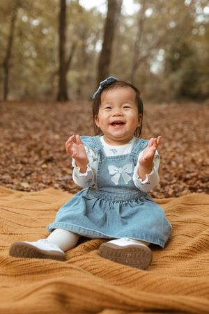 Baby wearing light blue denim dress sits on brown blanket surrounded by fallen autumn leaves.