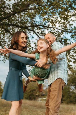 A family plays together outdoors on a sunny day, swinging and laughing in a grassy field under tree branches.