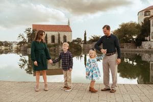 A family stands together on a stone walkway near a lake with a church in the background at sunset.