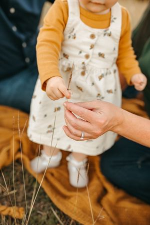 Small hands reach for dried grass stems while wearing a yellow sweater and patterned white top.