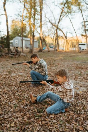 Two children in plaid shirts and jeans practice aiming toy rifles while crouching in autumn leaves at sunset.