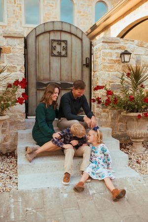 A family sits together on a stone porch step with red flowers and rustic wooden doors in the background.