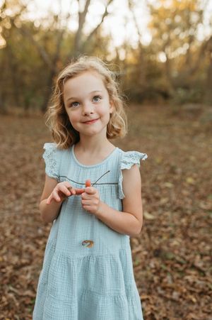 Child in light blue ruffled dress plays outdoors among autumn leaves in a forest setting.