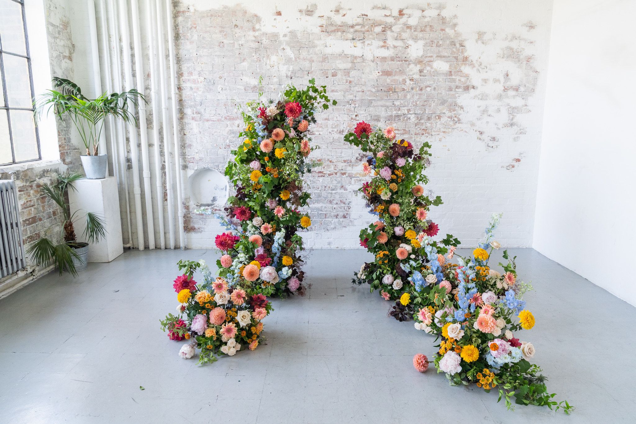 Two floral arrangements with colorful roses and greenery spiral upward against a white brick wall in an indoor setting.