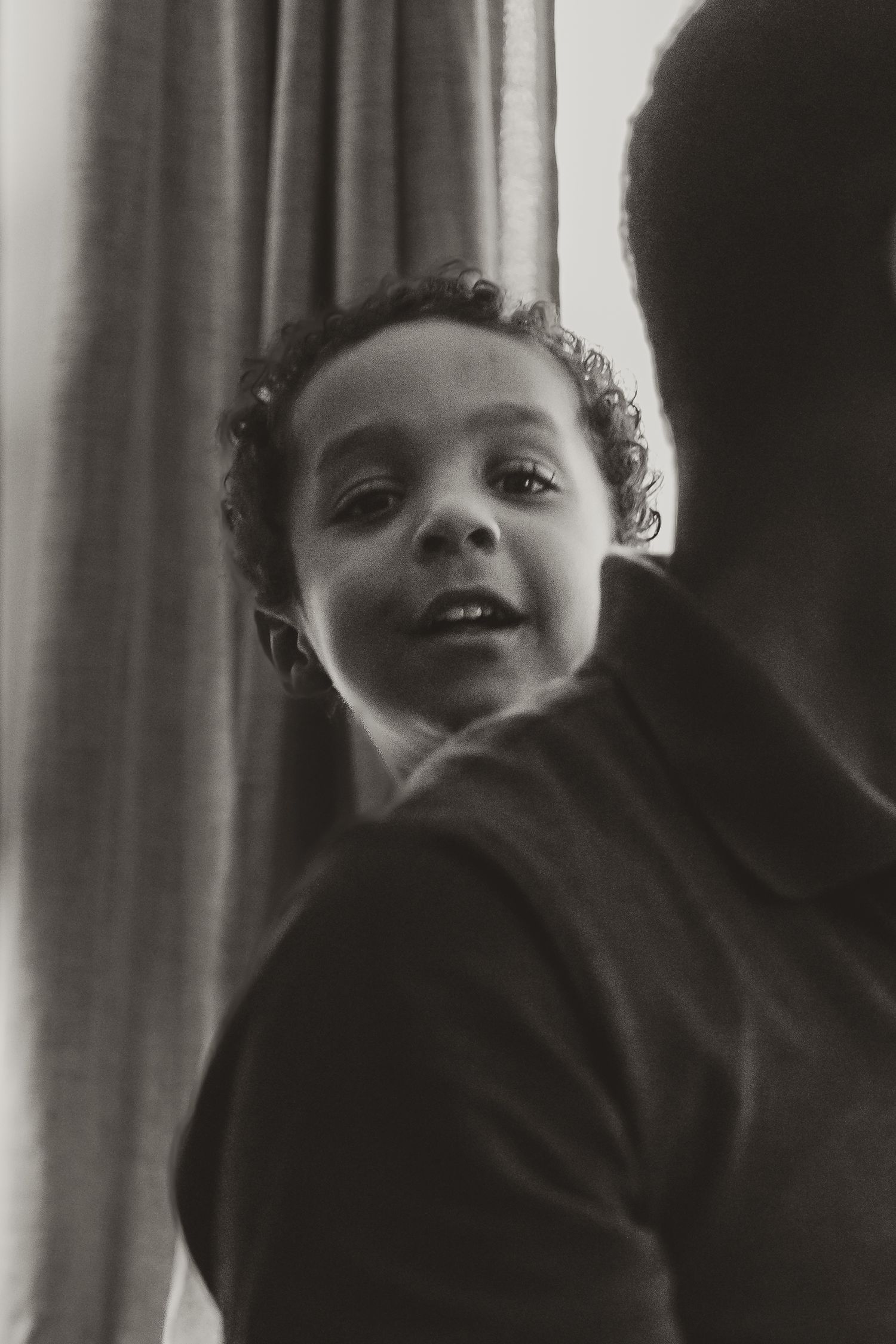 A black and white portrait photograph looking upward at a smiling child with curly hair against light curtains in the background.