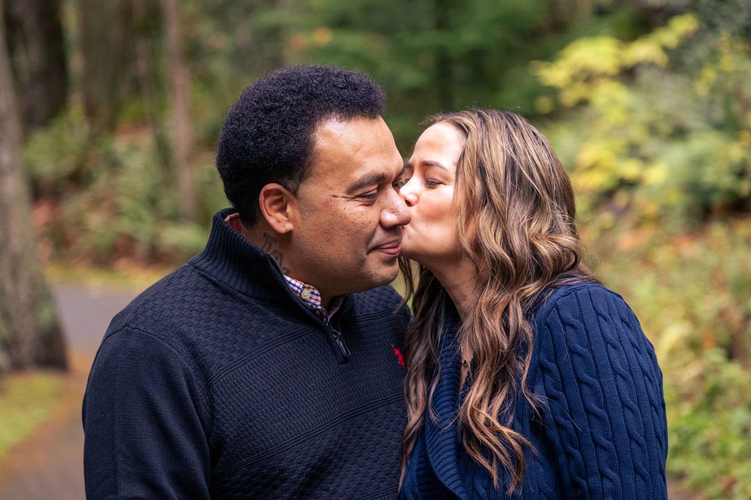 A woman kisses a man on the cheek as they stand together on a forest path, both wearing navy blue sweaters.