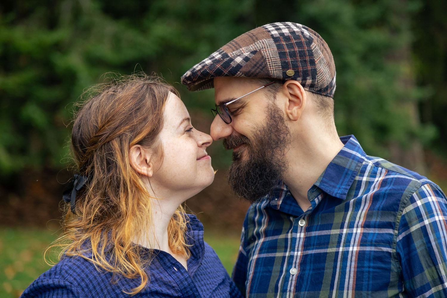 Couple in plaid shirts sharing a romantic moment outdoors, man wearing a flat cap and glasses, surrounded by green trees.