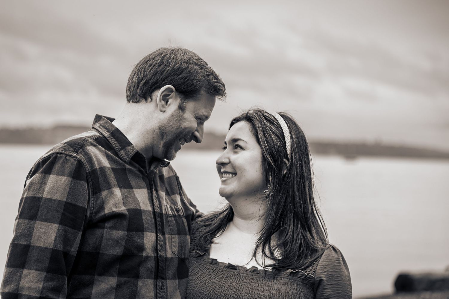 Black and white photo of a smiling couple gazing at each other outdoors near water.
