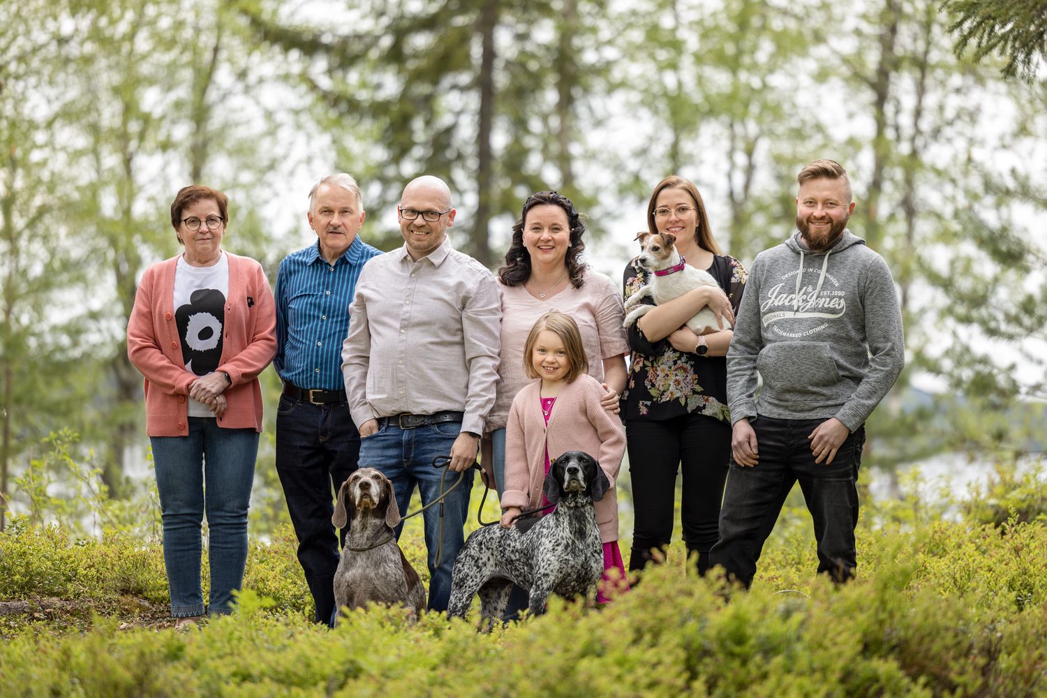Eri-ikäisiä ihmisiä metsä- ja järvimaisemassa mustikanvarpujen keskellä.