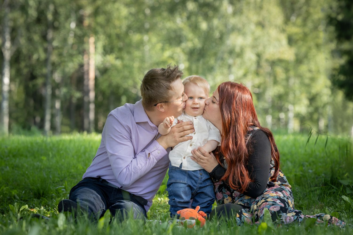 Vuoden ikäinen lapsi seisoo vanhempiensa välissä puistomaisema taustalla