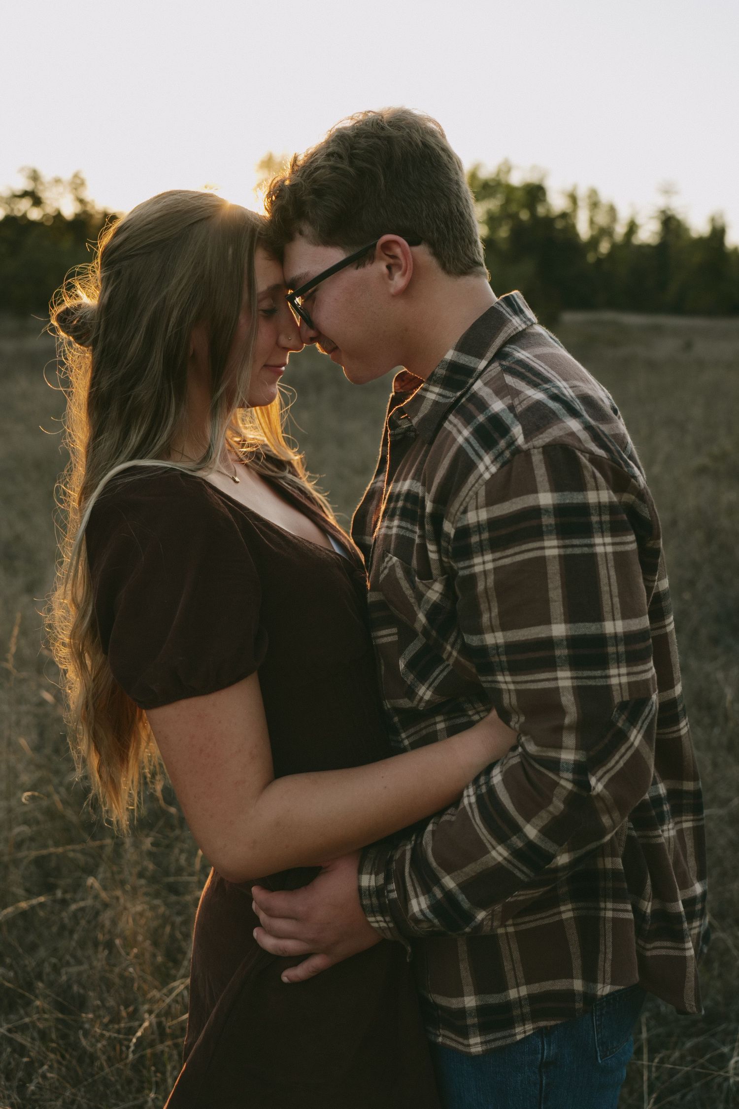 A romantic couple embraces in a golden sunset field while wearing casual attire.