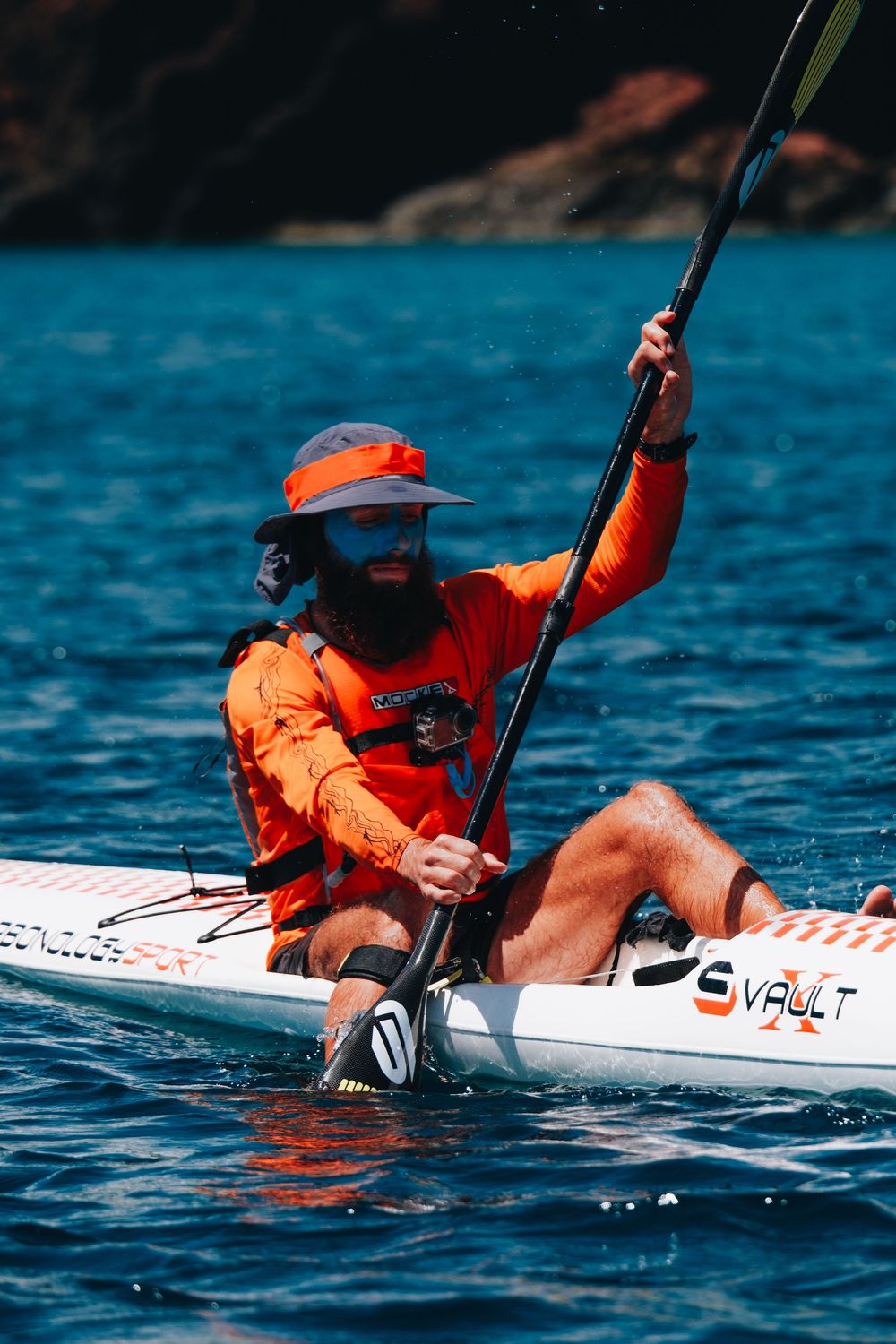A kayaker in bright orange safety gear paddling across blue water on a white vessel.