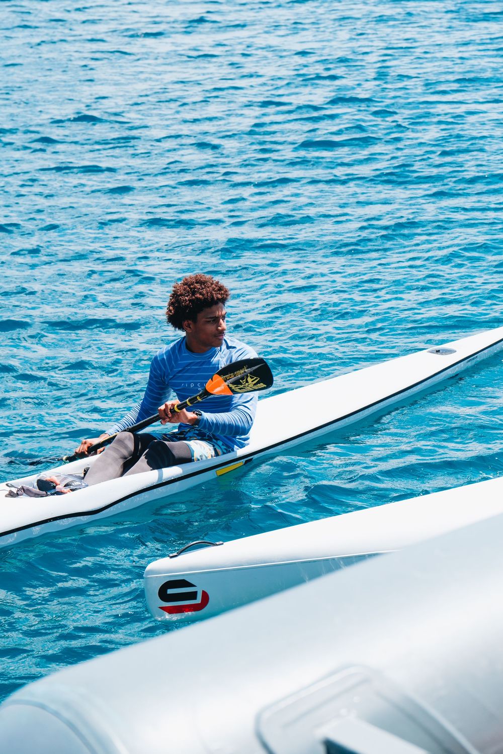 A person in a blue wetsuit sits aboard a sleek white water vessel on bright blue ocean waters during a sunny day.
