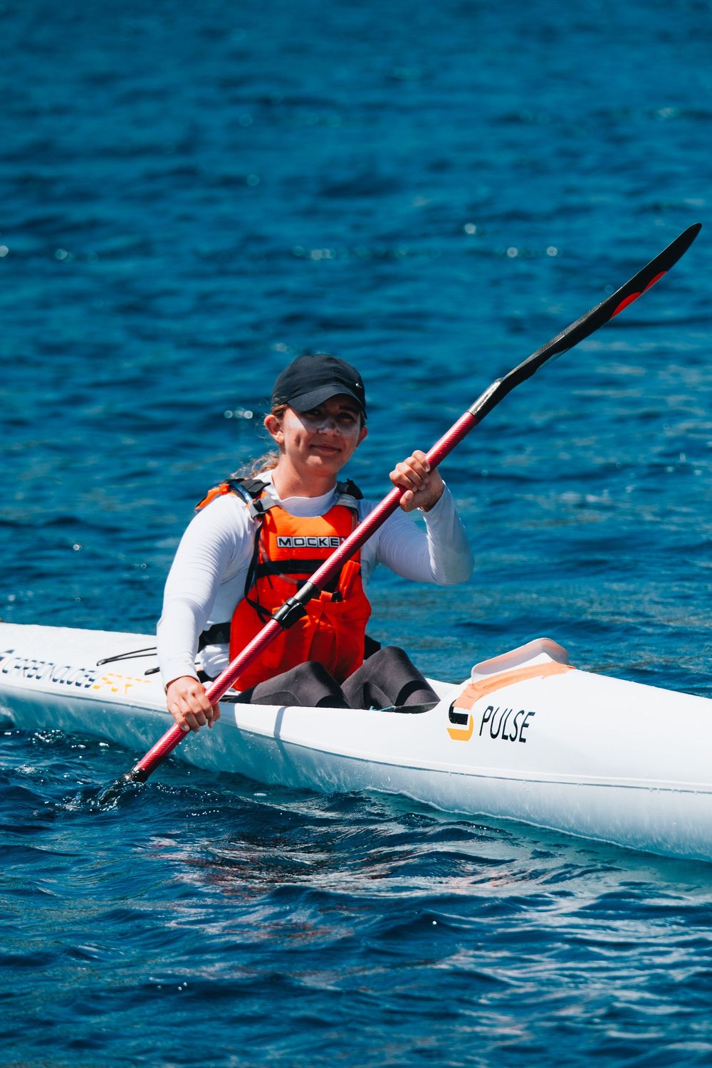 Athlete paddling a white racing kayak on bright blue water while wearing orange safety vest and black hat.