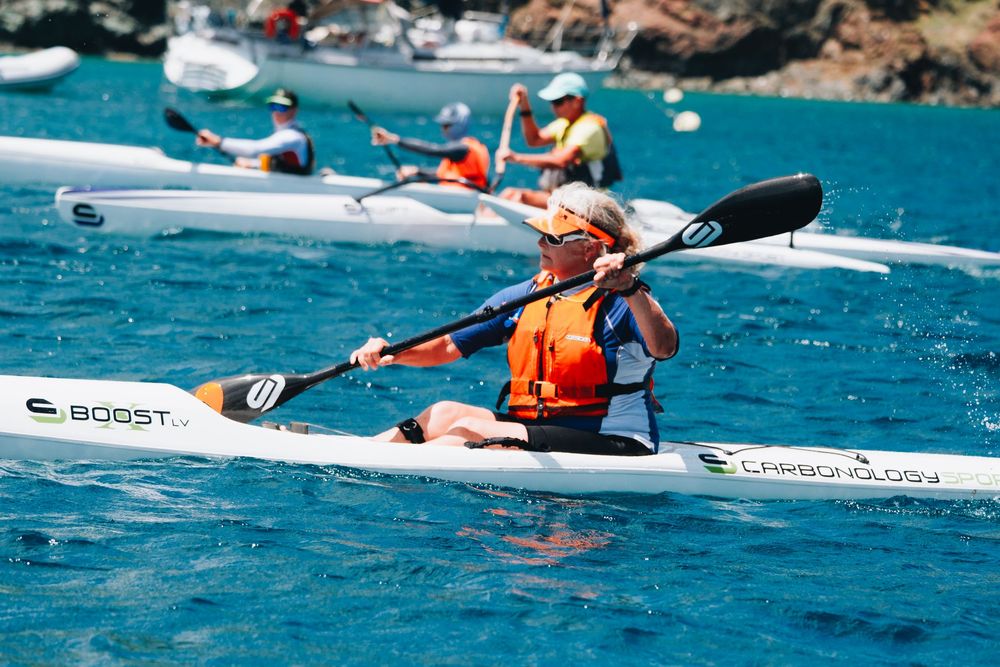 Kayaker wearing orange life vest paddling through bright blue ocean waters on a sunny day.