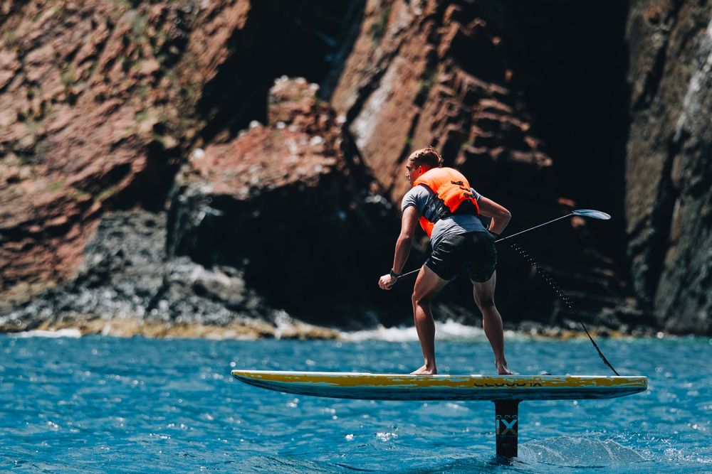 A person stand-up paddle boarding on bright blue waters near dramatic rocky cliffs performs a dynamic maneuver.