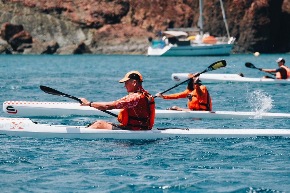 Two kayakers paddle through blue waters near red cliffs with boats in the background.