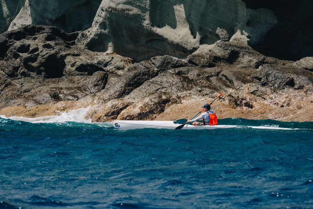 A kayaker in red paddles along dramatic coastal cliffs with bright blue ocean waters below.