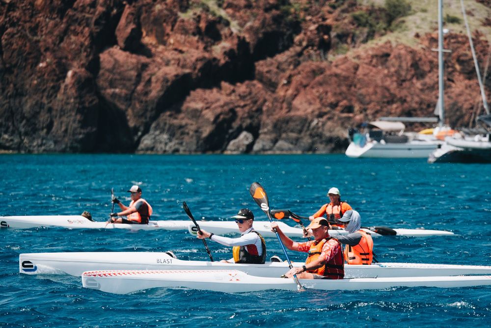 Kayakers paddling through blue waters near red rocky cliffs with a white sailboat anchored in the background.