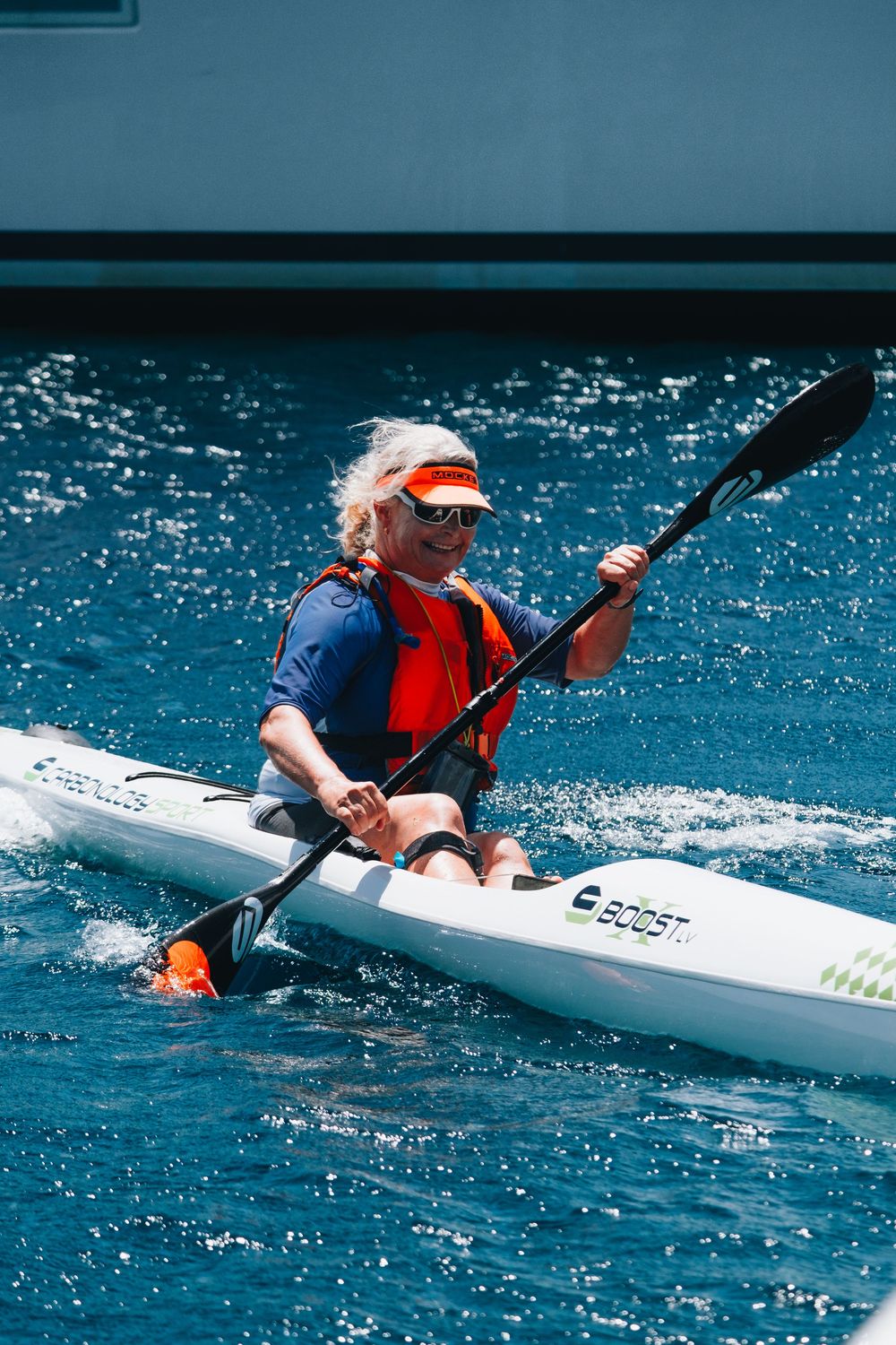 Kayaker wearing an orange life vest and navy shirt paddles through bright blue ocean waters on a white kayak.