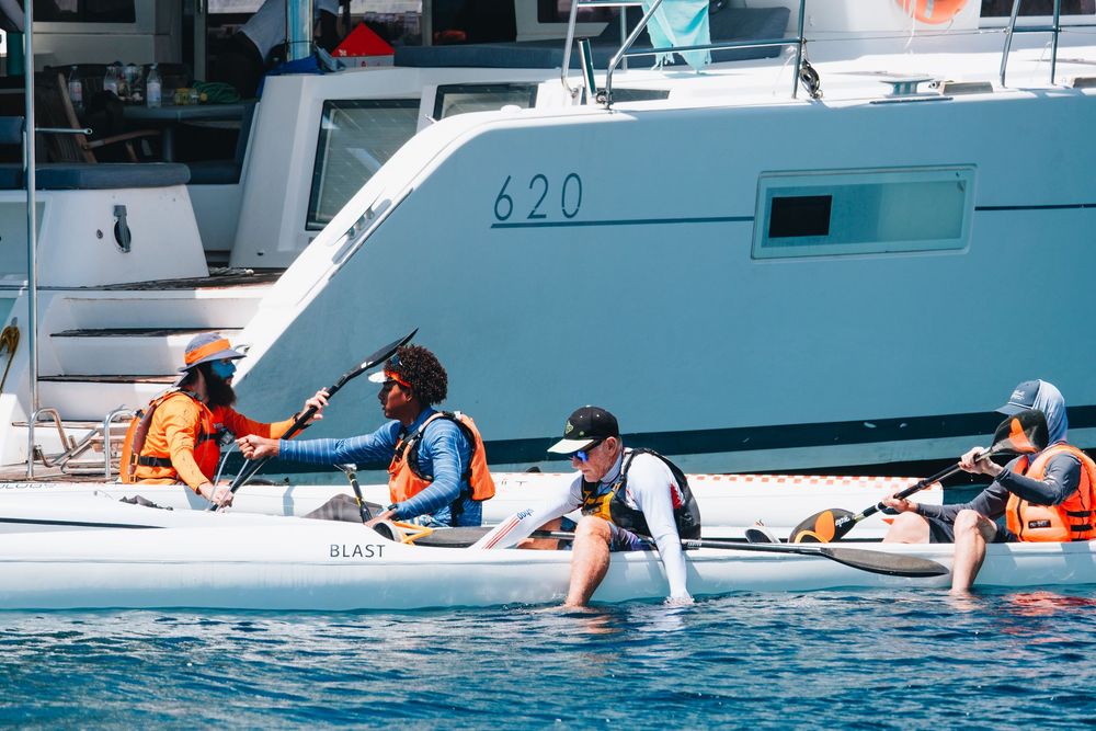 A group of people in orange life jackets gather on a small boat near a large yacht in blue waters.