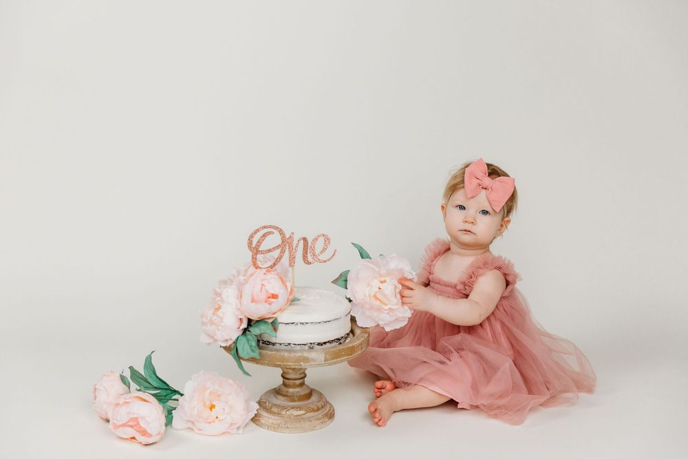 First birthday setup with baby in dusty rose tulle dress next to white cake and pink flowers.