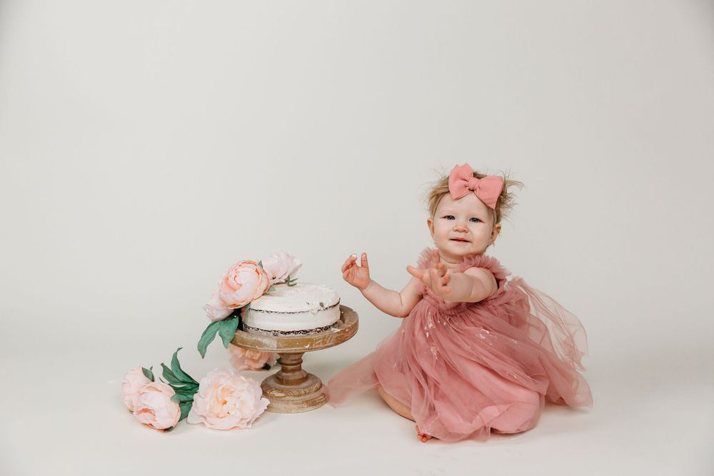 Baby in dusty rose tulle dress sitting beside a cake stand decorated with pink flowers.
