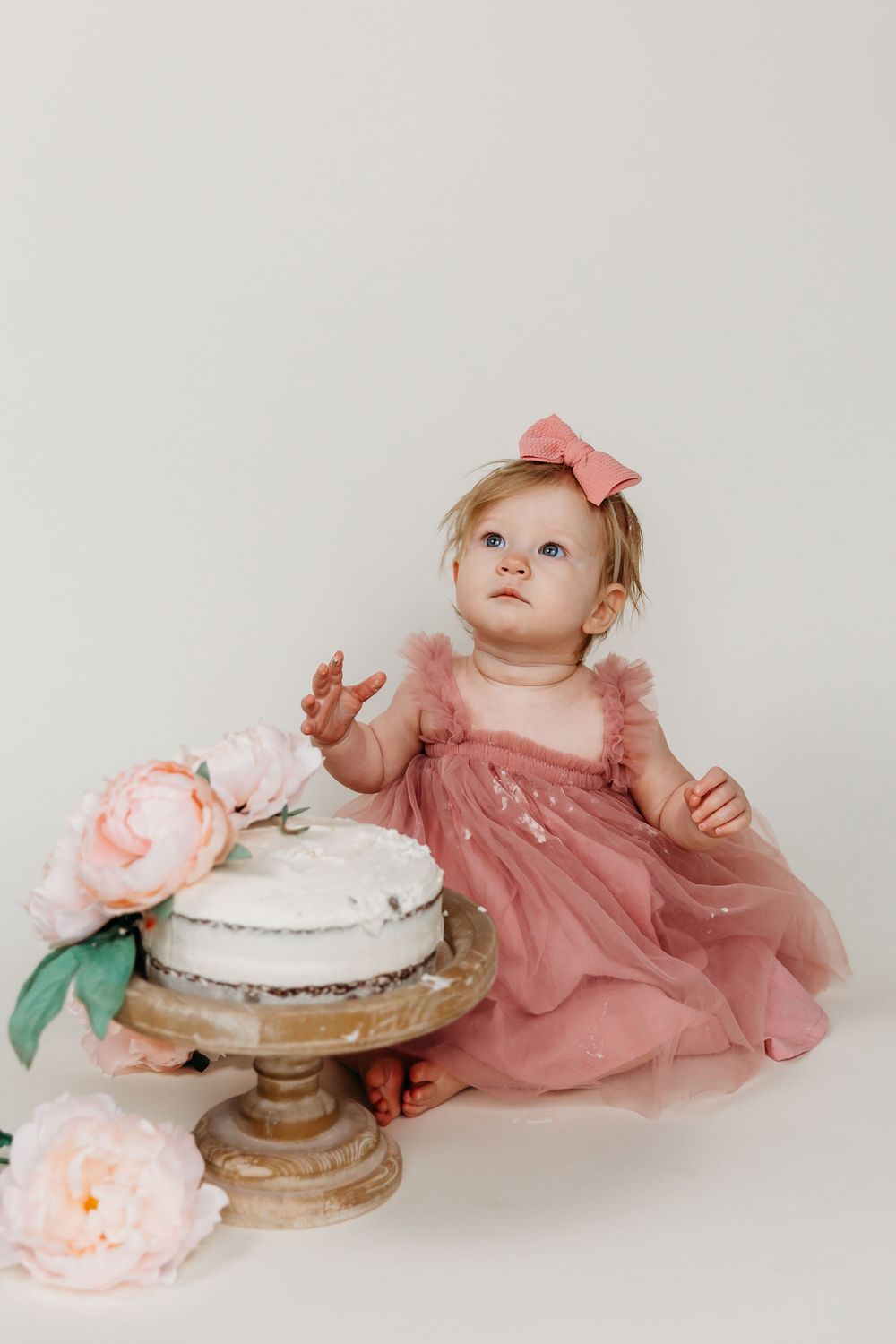 Baby wearing pink dress reaching toward white cake on vintage pedestal with floral decorations.