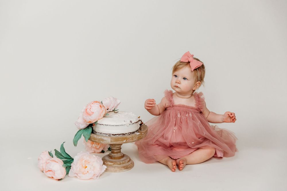 Baby in pink dress sitting beside decorated cake stand with pink roses on white background.