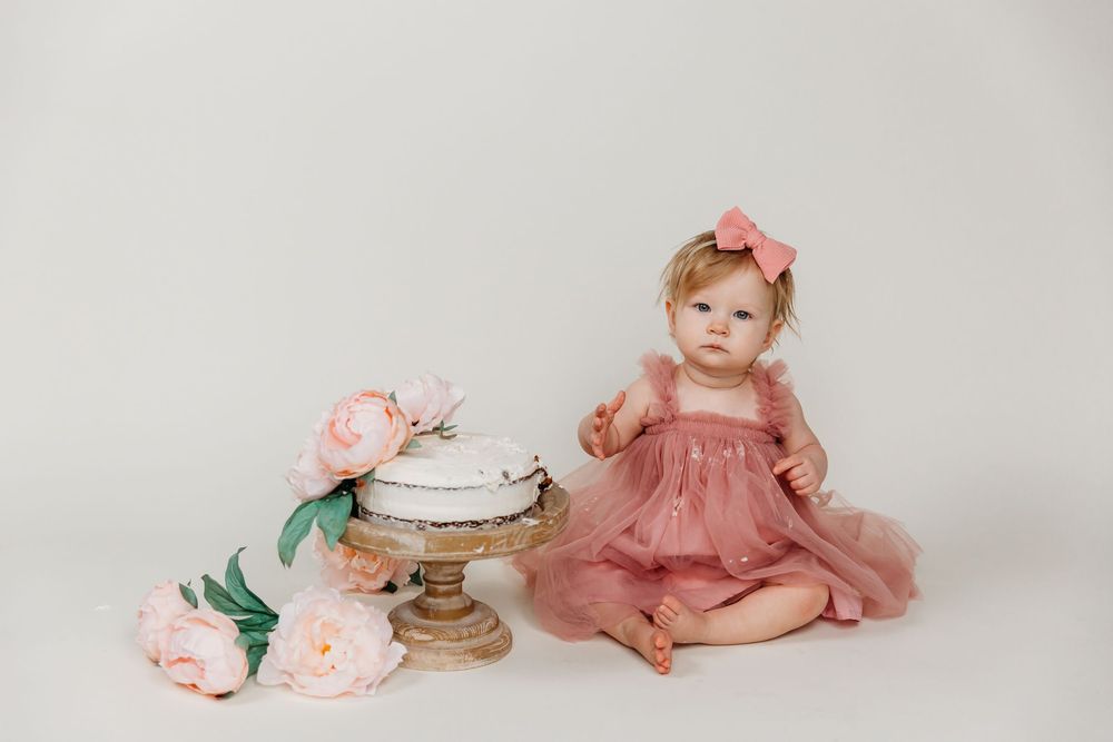 Baby in pink tulle dress reaching for cake on rustic stand with floral accents.