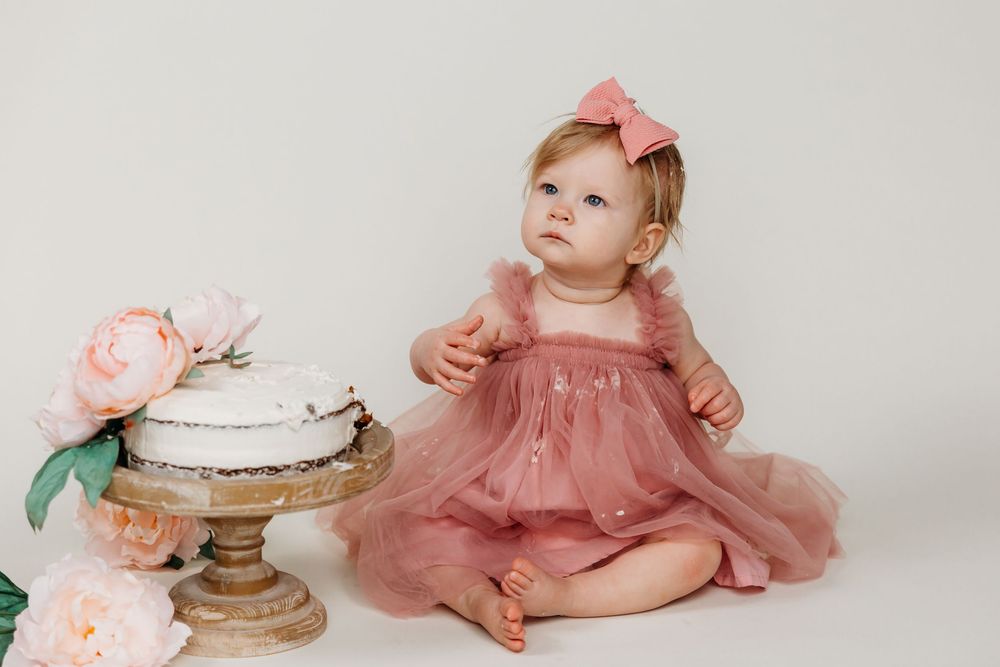 Baby in dusty rose dress looking up at cake on vintage stand with pink flowers.
