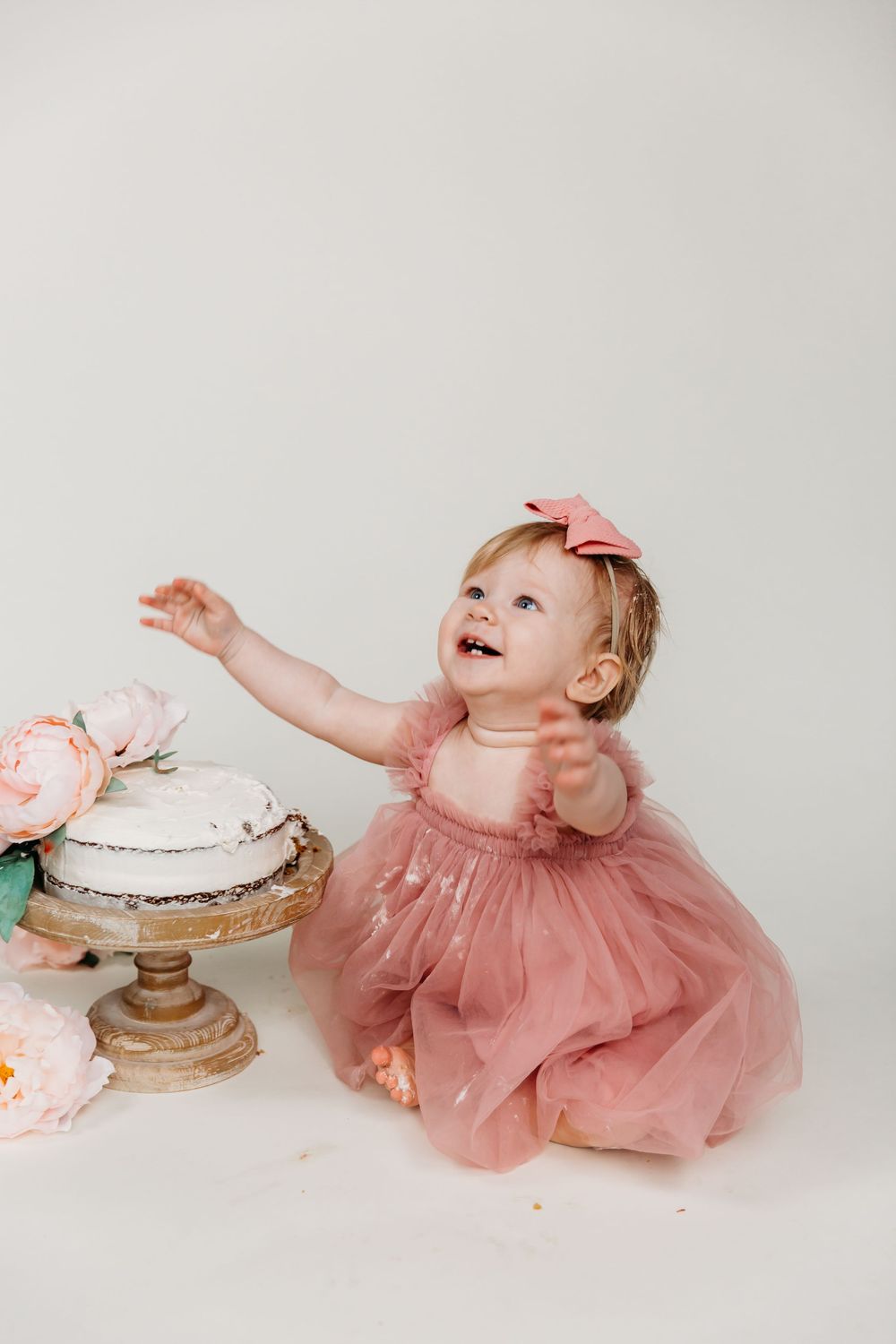 Baby in pink dress reaching upward during cake smash photo session with floral decorations.