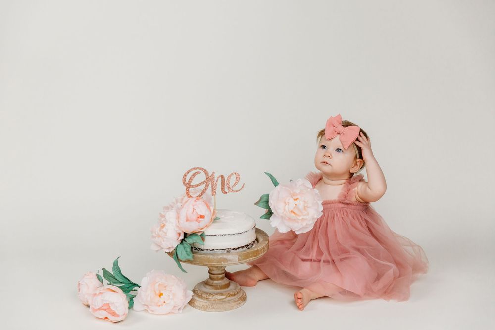 Baby in pink tulle dress reaching toward elegant white birthday cake with floral decorations.