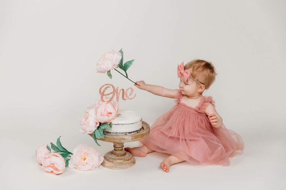Baby in pink dress reaching for flowers beside white birthday cake on vintage stand.