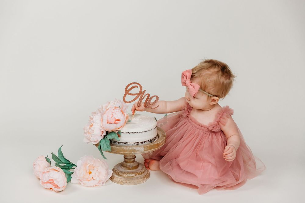Baby in pink dress reaching for elegant white cake with 'One' topper and roses.