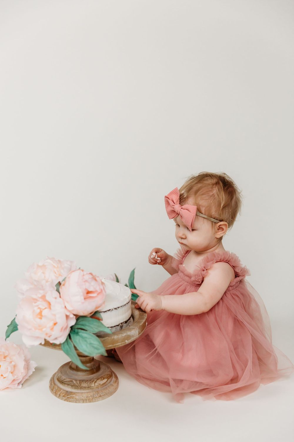 Baby in tulle dress exploring birthday cake setup with pink peonies.