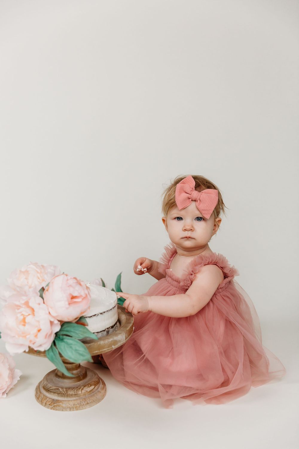 Baby in pink dress sitting beside vintage cake stand with floral decorations.