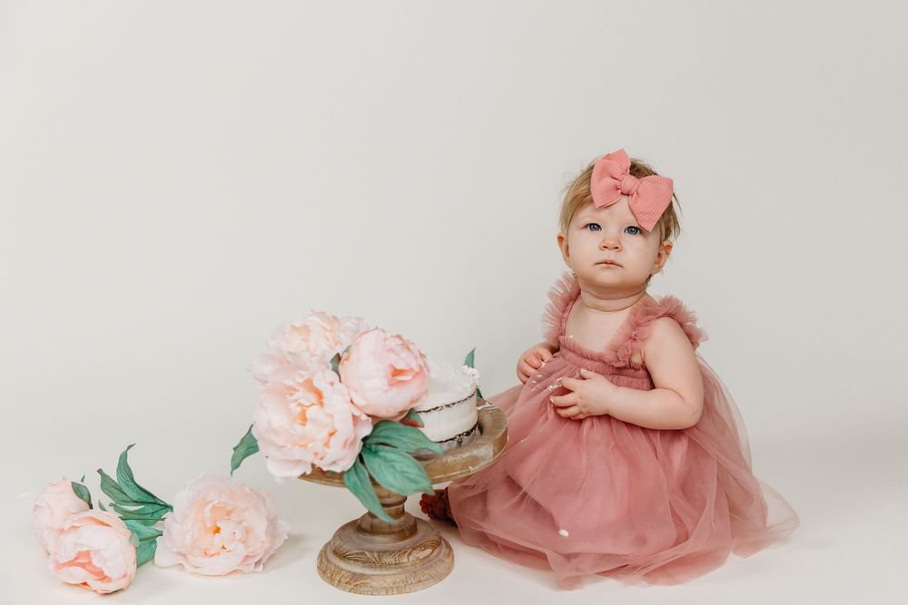 A cake smash photo setup with pink roses and a rustic cake stand against a white backdrop.