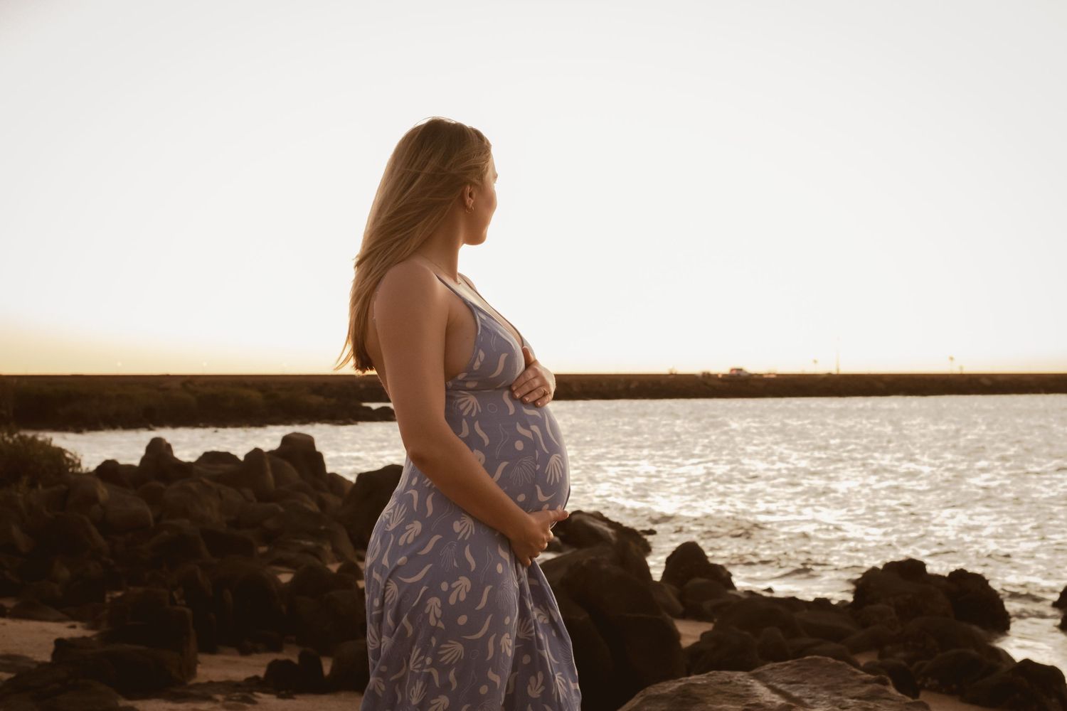 Woman in a floral dress standing on rocky coastline during sunset.