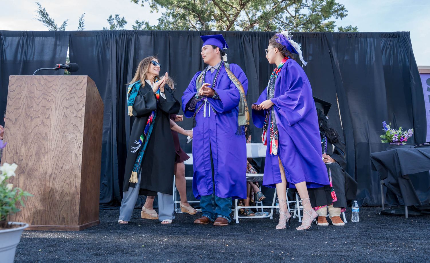 Students in blue graduation gowns converse with faculty member during outdoor commencement ceremony.