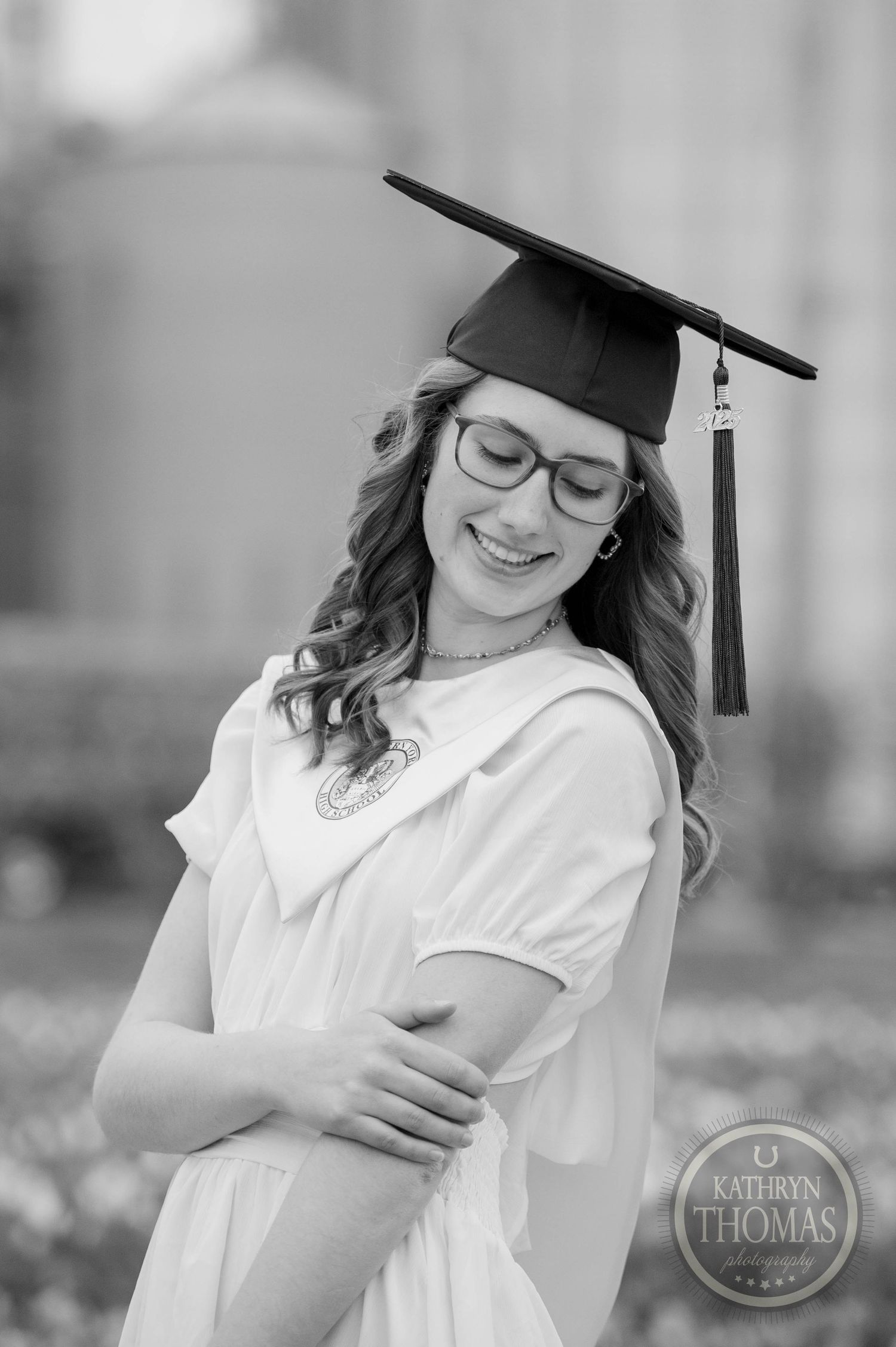Black and white graduation portrait showing a graduate in a cap and gown with glasses posing outdoors.