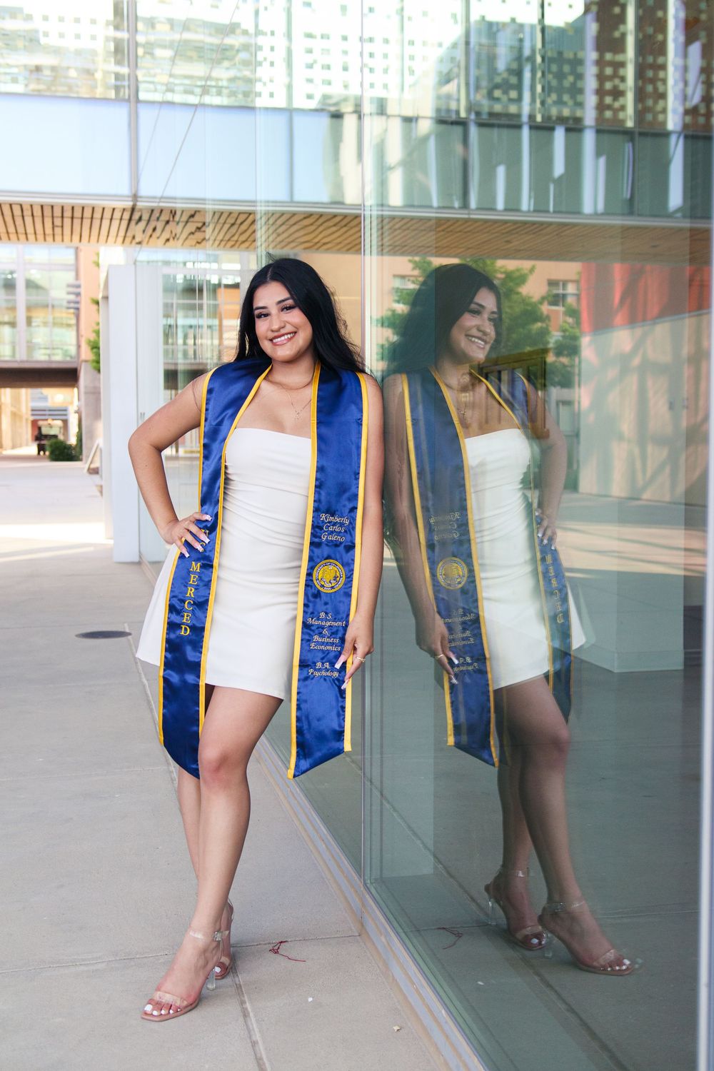 Smiling graduate in white dress with blue and gold graduation sash outdoors.