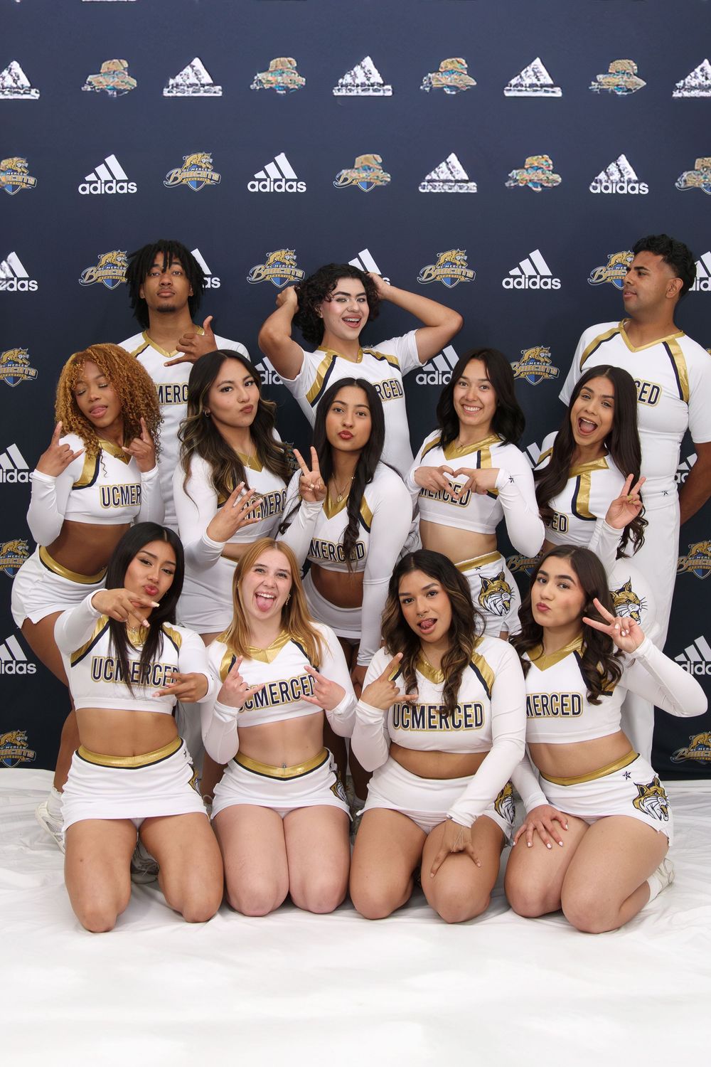 UC Merced cheerleading squad posing together in white and gold uniforms in front of an Adidas branded backdrop.