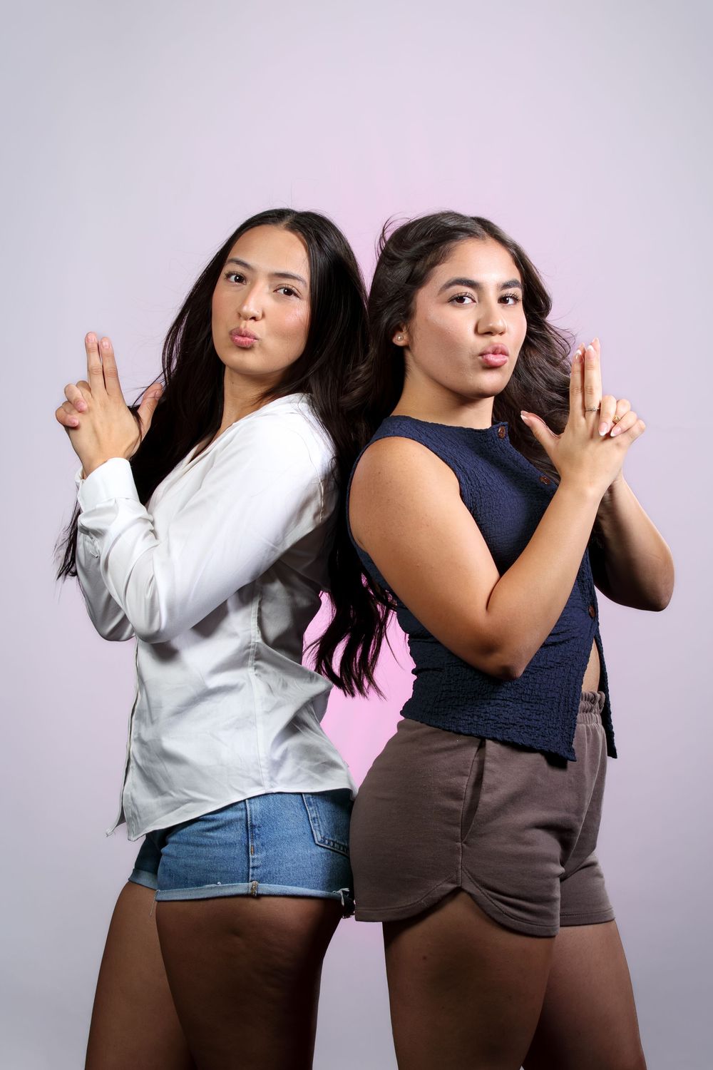 Two young women posing back-to-back making finger gun gestures on pink background.