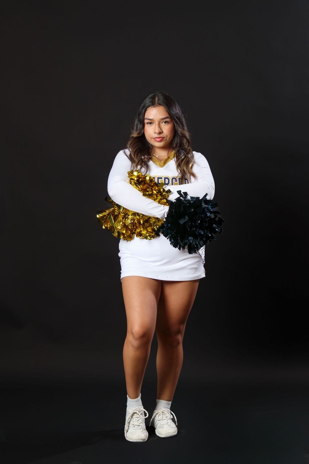 A cheerleader in white and gold uniform poses with pom-poms against a dark background.