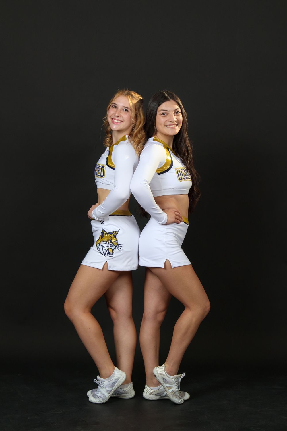 Two cheerleaders in white UCMERCED uniforms pose back-to-back against a dark background.