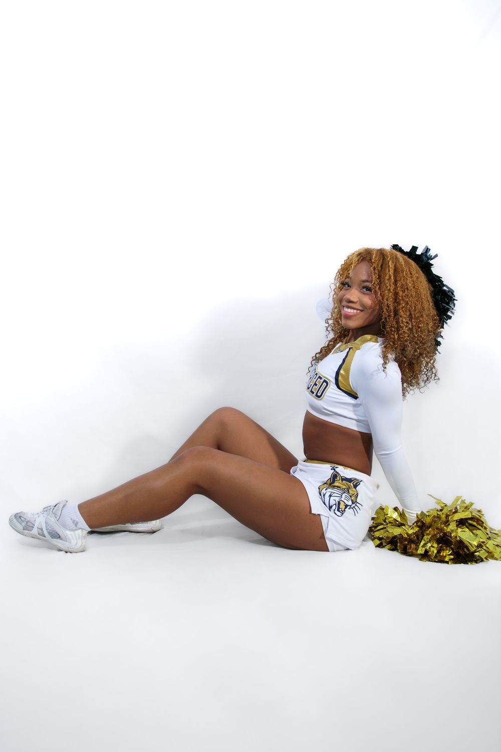 A smiling UC Merced cheerleader in uniform sitting on a white floor holding gold pom-poms with a hair bow.