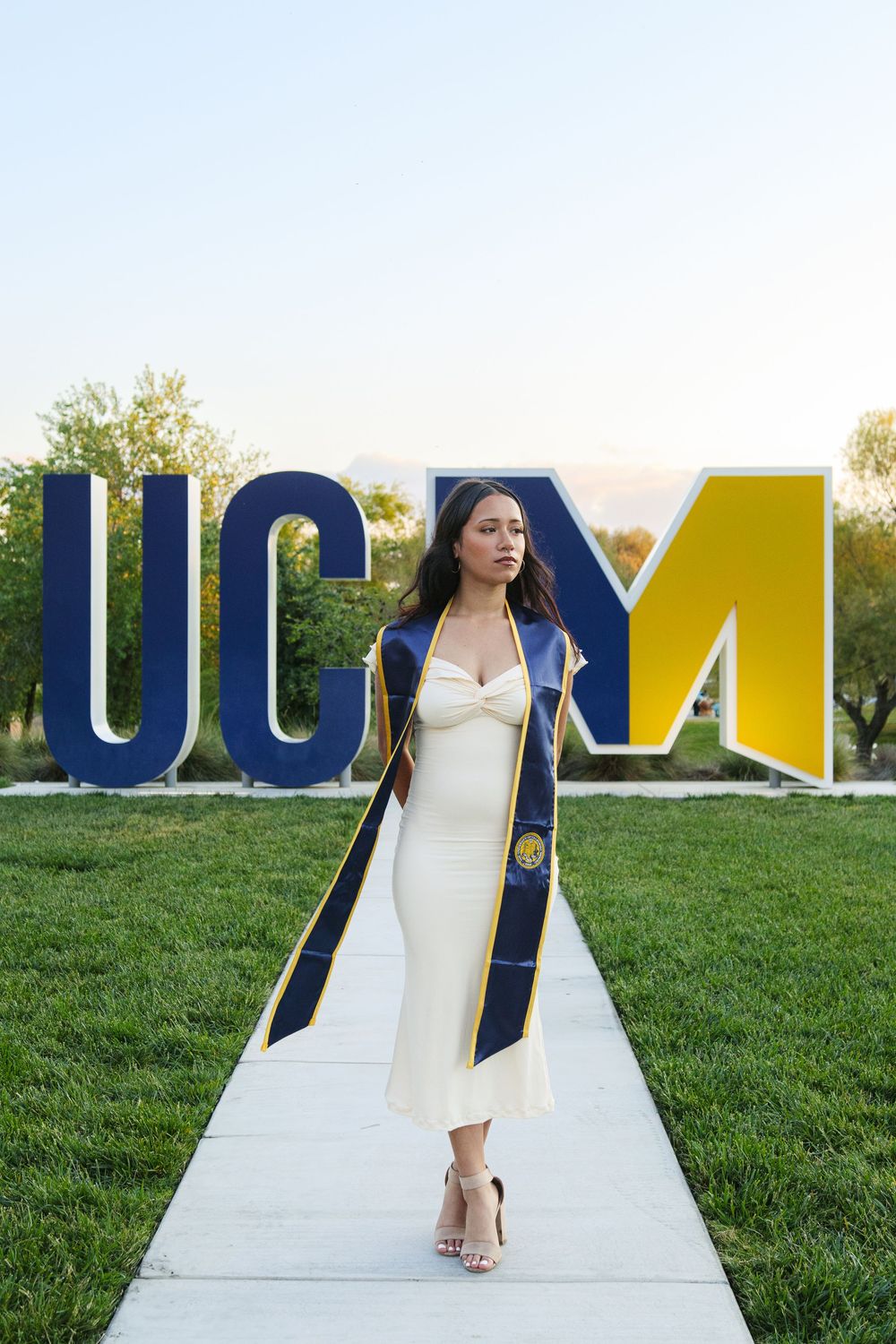 Female graduate in white dress with UC Merced stole walks confidently before large UCM sign outdoors.