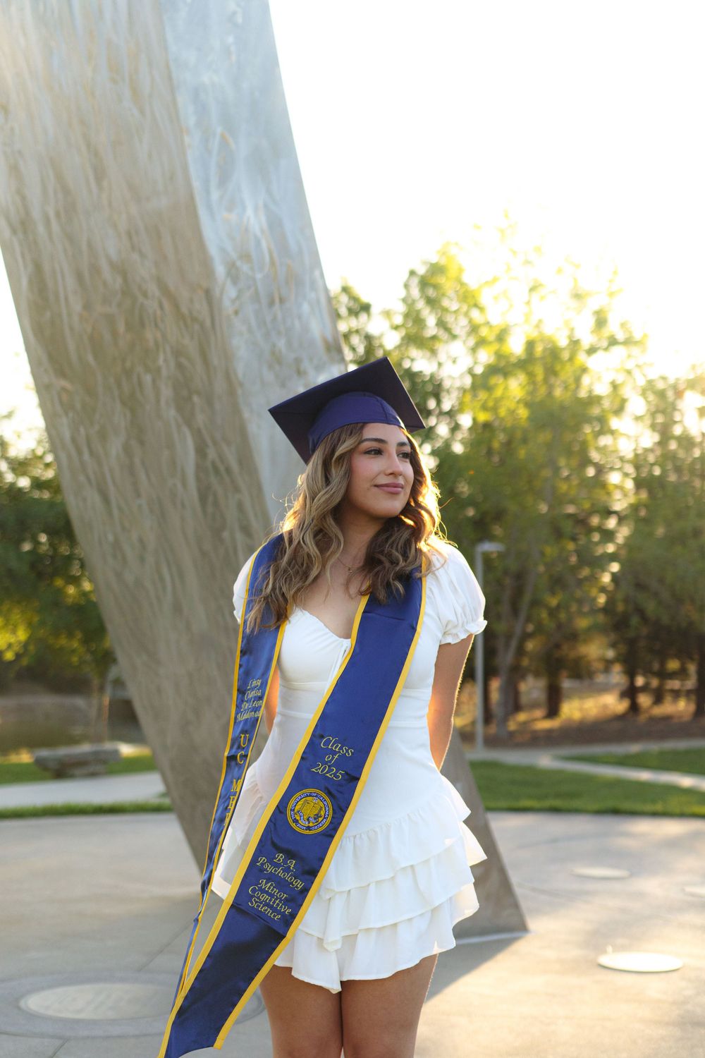 Female graduate in white dress and cap poses with UC Merced sash near iconic campus sculpture.