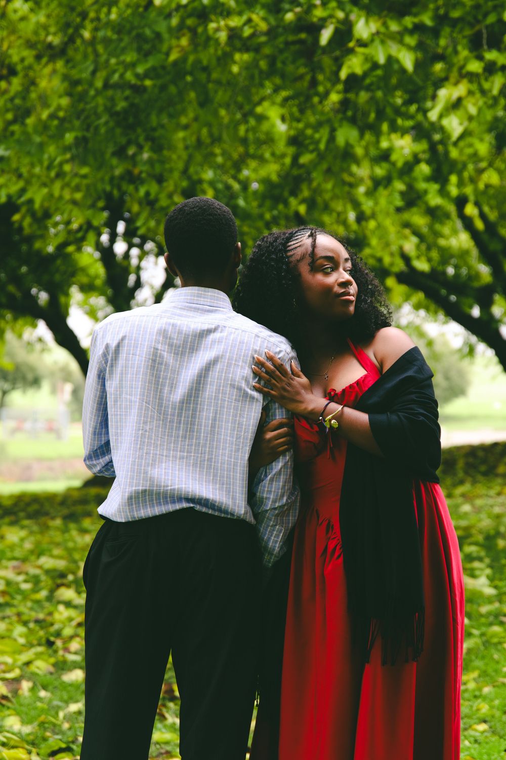 A couple embraces tenderly outdoors, with the woman in a red dress leaning against the man.